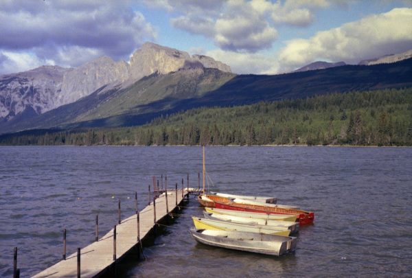Bowfort Lake in the foreground with the vertical cliffs of Yamnuska rising in the distance. The dock juts out in the lake with several row boats and a single sailboat tied up to the right of the dock and streaming out on the painters, down wind. The lake here is relatively calm not like the waves described in the story.